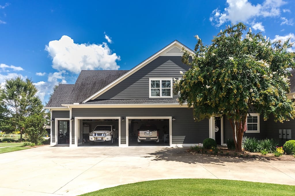 The side view of a large gray craftsman new construction house with a landscaped yard a three car garage and driveway with two doors open.