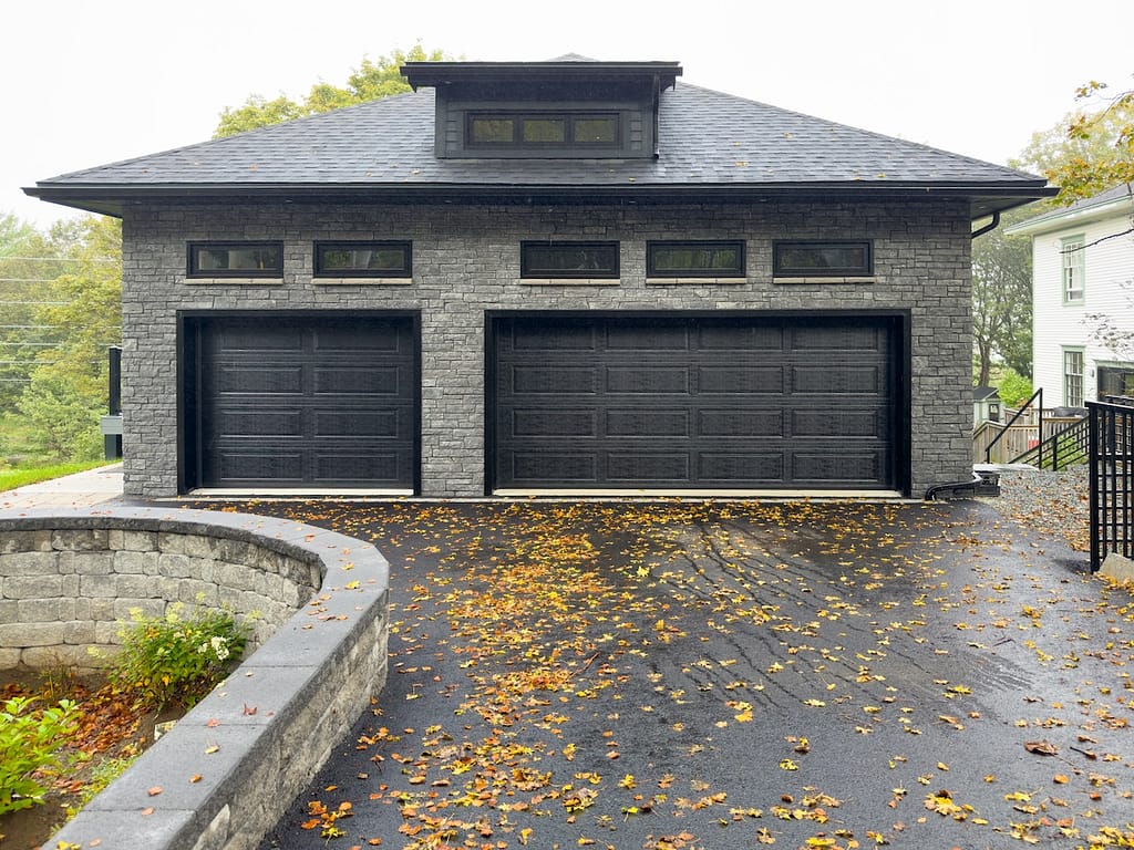 A double and single-width black metal garage door with panels in a grey brick luxurious residential outcrop building. There are small glass windows above the doors with a dormer window on the roof.